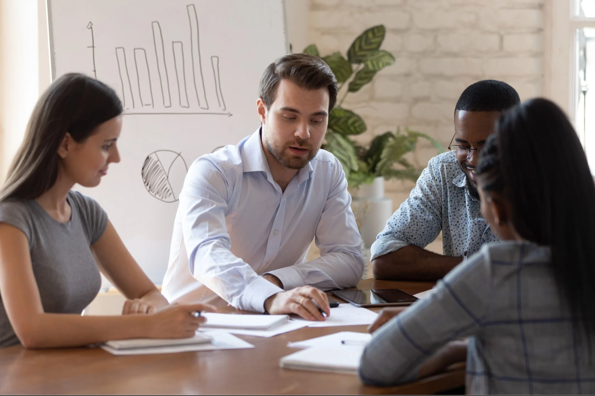 Team meeting around a wooden table with a strategy chart on the whiteboard, reviewing plans together